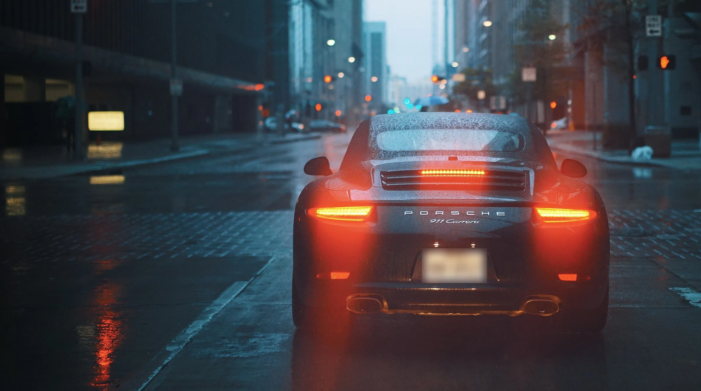 Porsche 911 Carrera preto visto de trás em rua molhada ao entardecer, com lanternas acesas