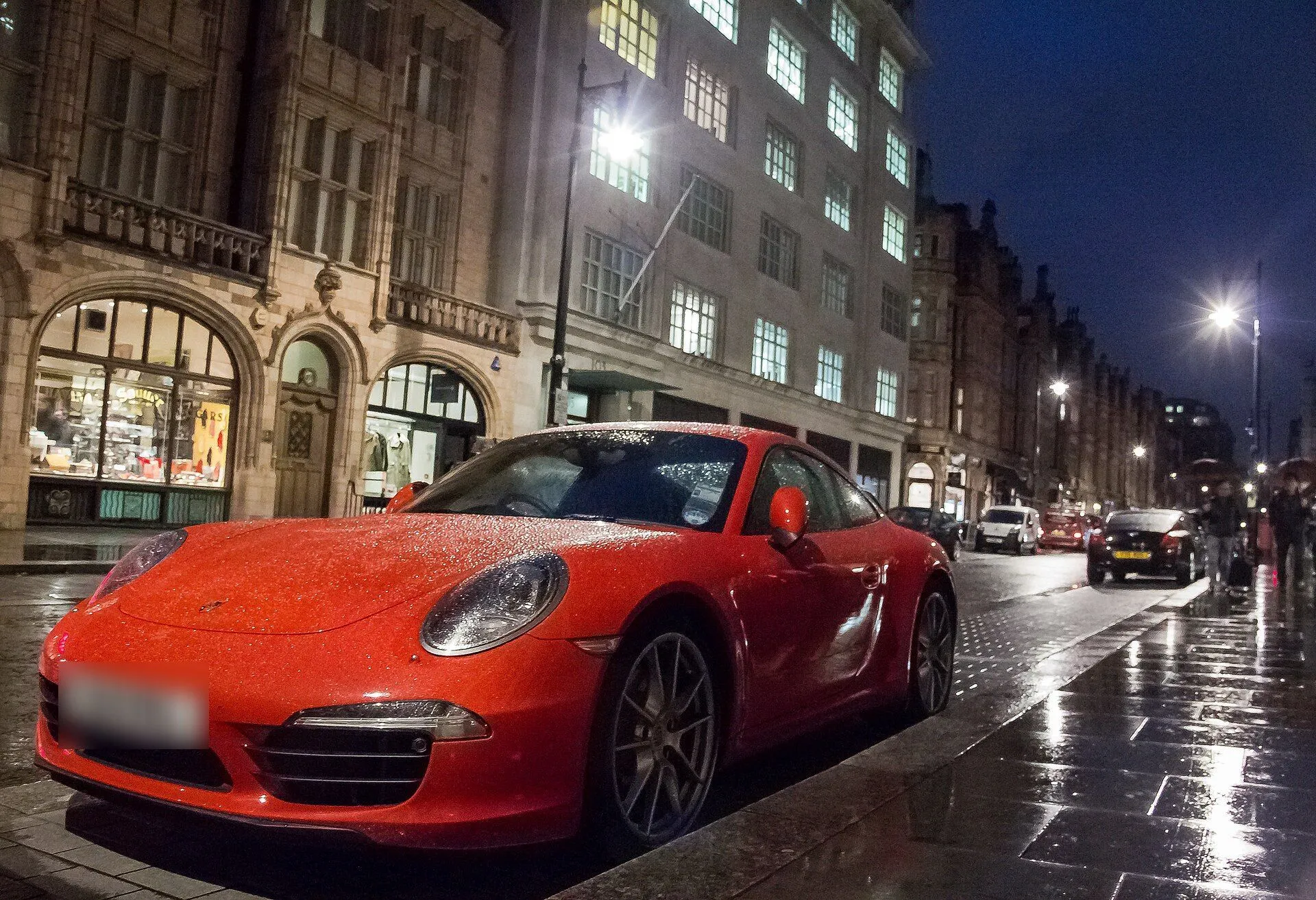 Porsche 911 vermelho estacionado em rua molhada à noite, com prédios iluminados ao fundo